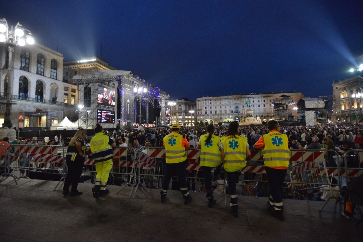 Cinque soccorritori in divisa gialla disposti in linea di spalle davanti alle transenne durante un evento musicale affollato in piazza di notte.