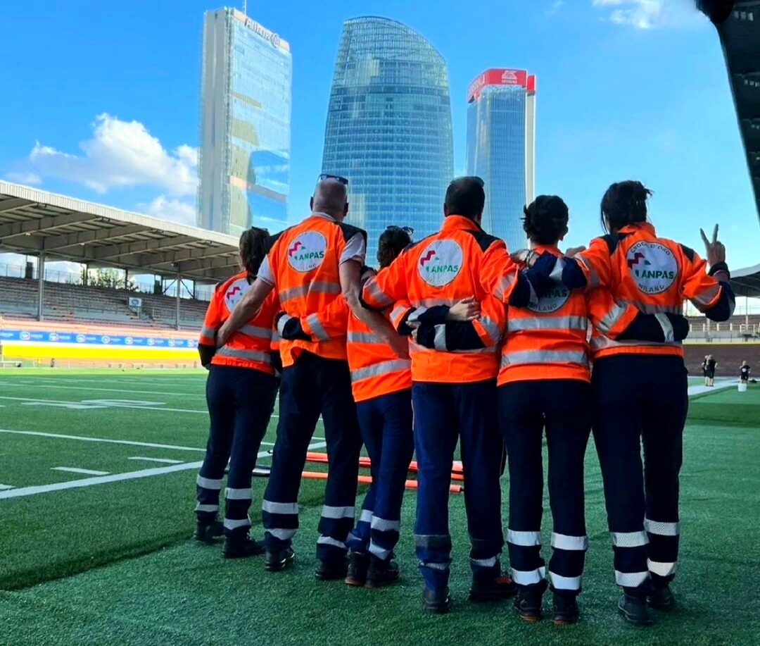 Vista di spalle di un gruppo di soccorritori abbracciati che osservano i grattacieli di Milano durante un servizio di assistenza eventi.