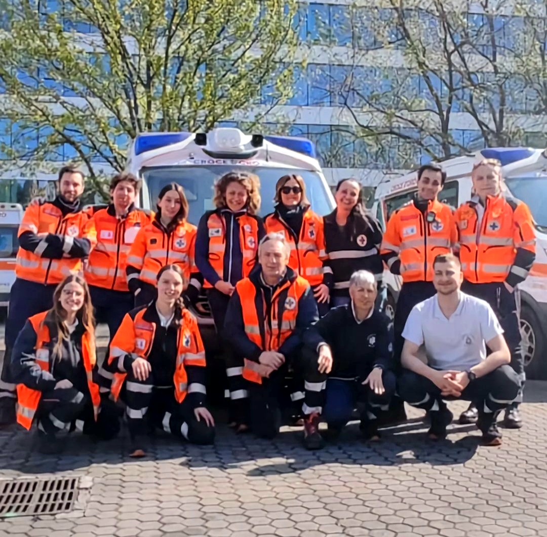 Un grande gruppo di volontari e soccorritori della Pubblica Assistenza in divisa ufficiale arancione ad alta visibilità posa sorridente all'aperto.