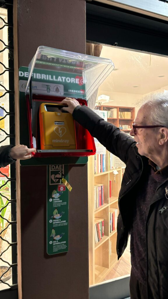 Un cittadino anziano mentre si avvicina alla teca del defibrillatore pubblico DAE Forlanini per aprirla, in un contesto urbano.