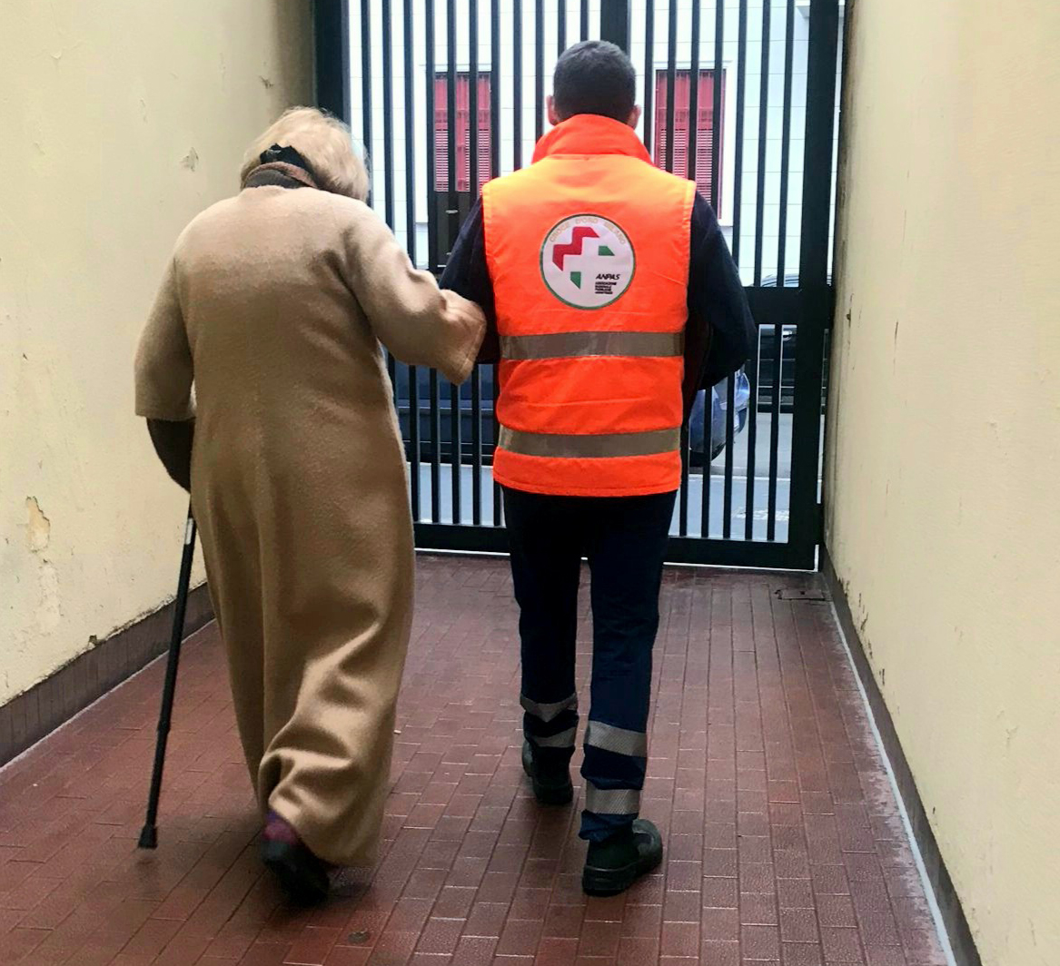 Un volontario della Pubblica Assistenza cammina a braccetto con una signora anziana per assisterla e accompagnarla in sicurezza verso l'ingresso di una struttura.