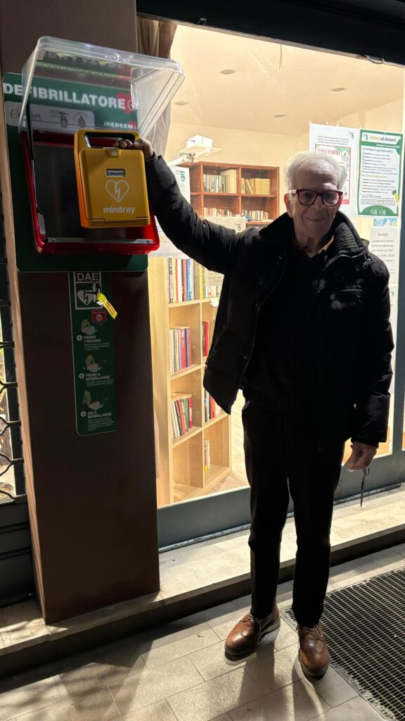Un cittadino in giacca scura posa accanto al totem del defibrillatore PAD Forlanini a Milano indicando la teca salvavita.