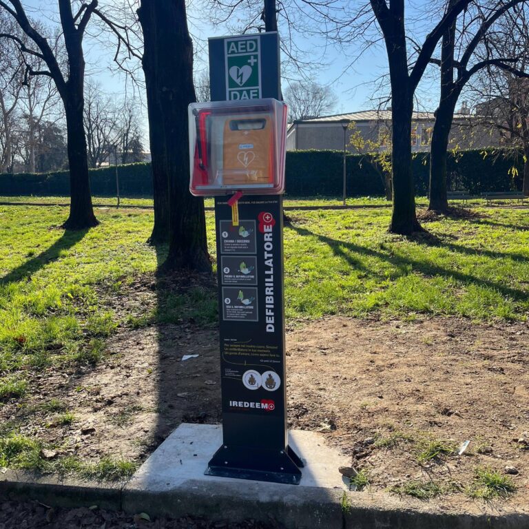 Primo piano del totem PAD Martinengo con teca protettiva per defibrillatore automatico in un'area verde a Milano.
