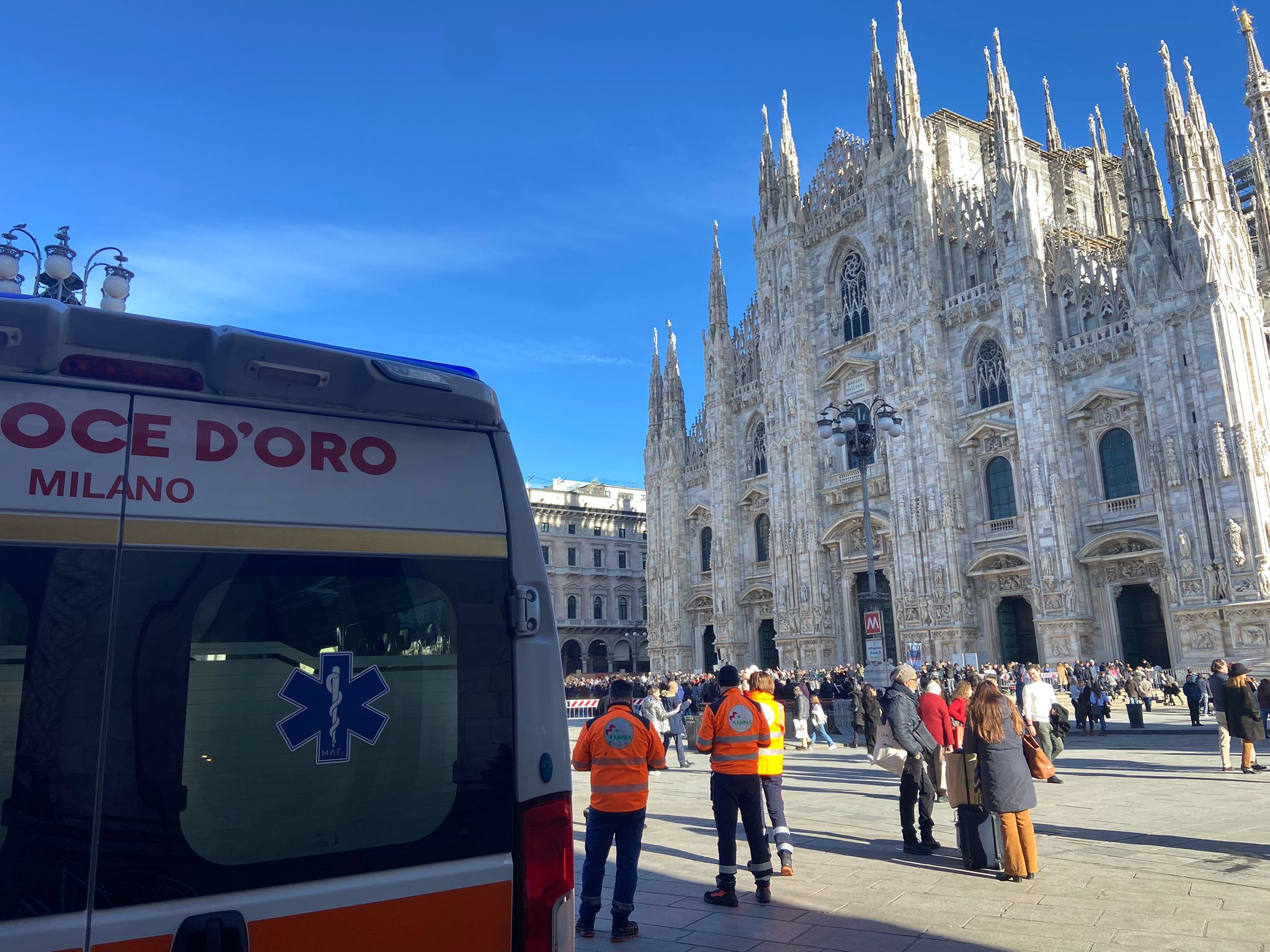 Vista parziale del retro di un'ambulanza della Croce d'Oro e soccorritori in divisa arancione in Piazza del Duomo a Milano, con la facciata del Duomo sullo sfondo.