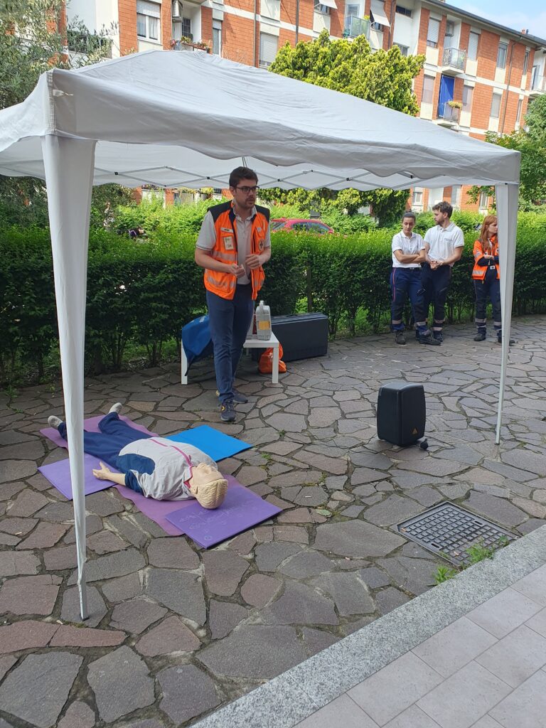 Un istruttore in piedi sotto il gazebo bianco spiega le linee guida internazionali sull'uso del DAE al pubblico di CentoPassiPerLaVita.