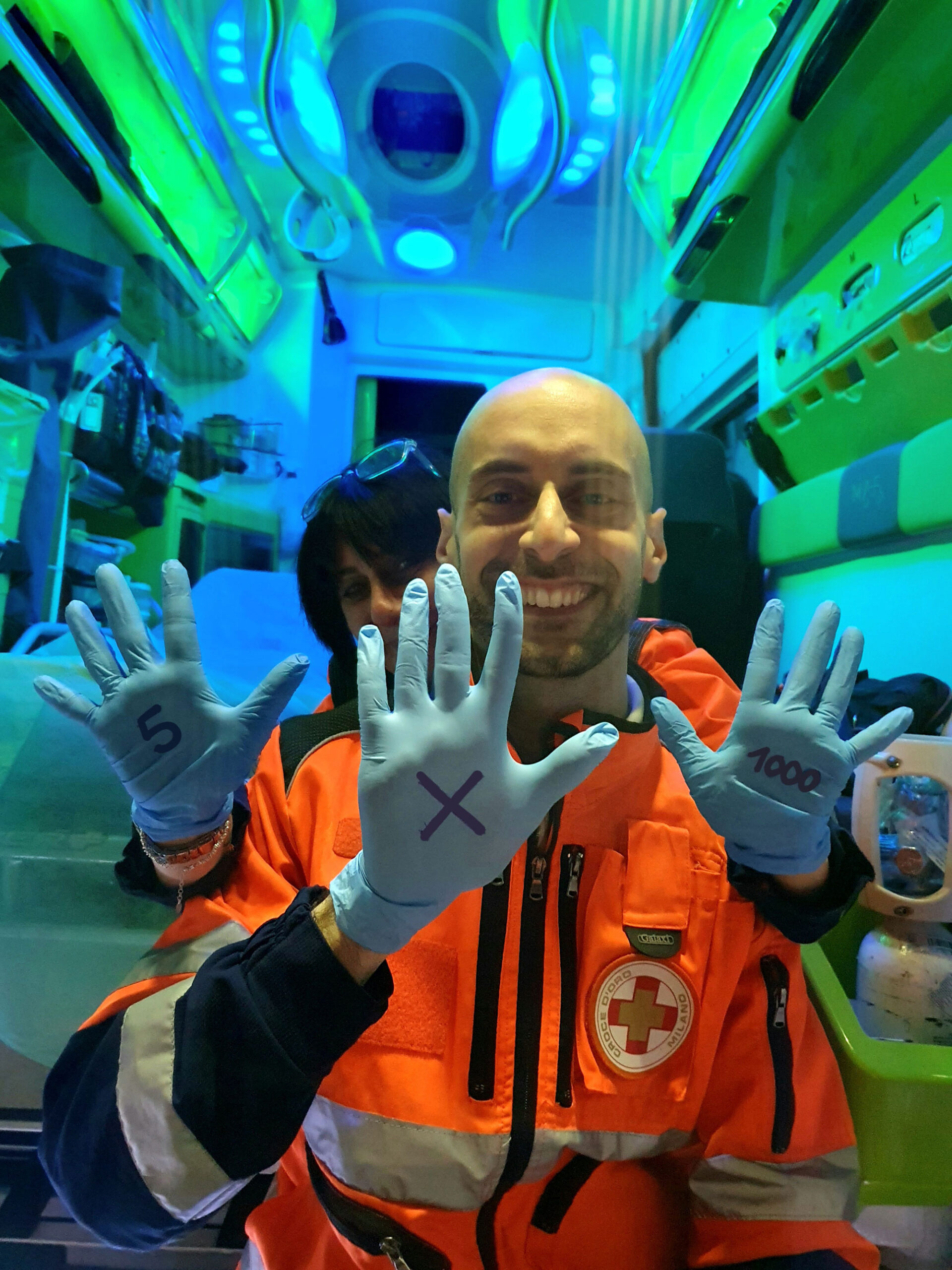 Un volontario sorridente in divisa arancione della Croce d'Oro mostra i palmi delle mani protetti da guanti azzurri per la campagna del 5x1000.
