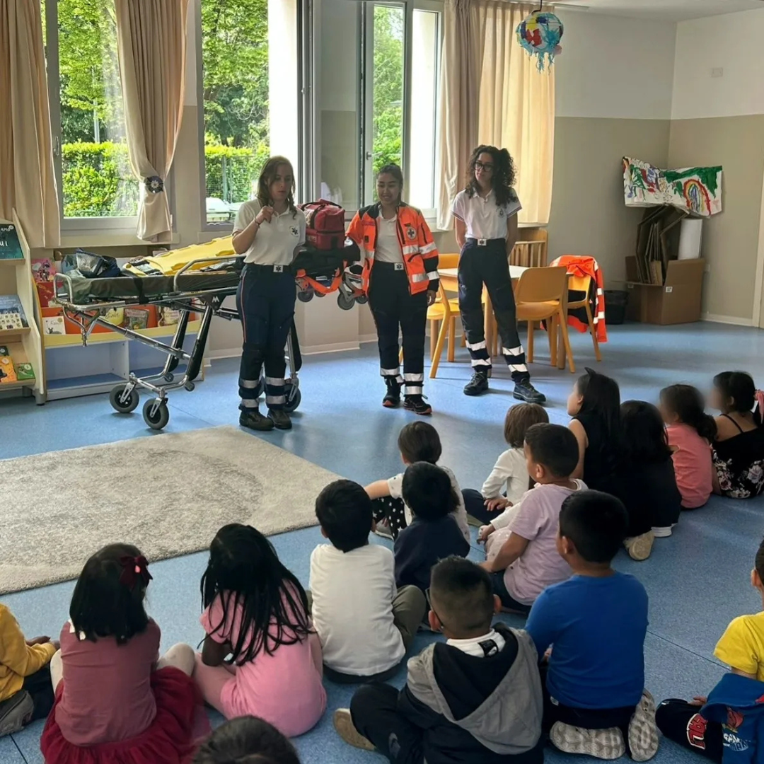 Operatori della Croce d'Oro Milano mostrano un'ambulanza e una barella a un gruppo di bambini seduti durante un incontro didattico di sensibilizzazione.