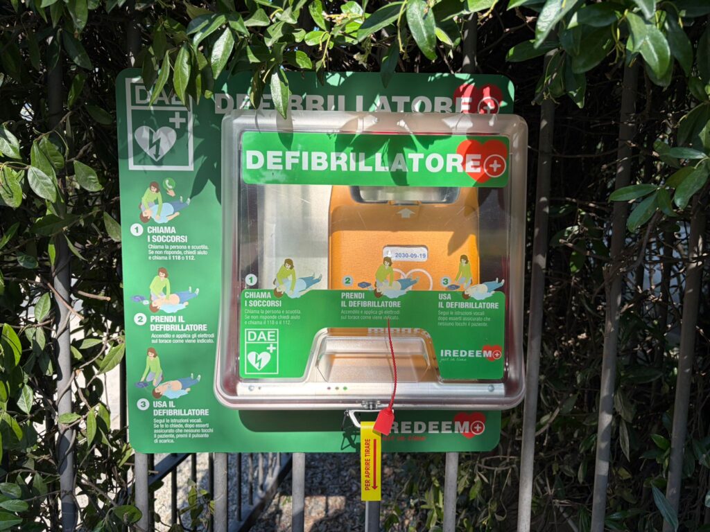 Teca verde per il posizionamento pubblico di un defibrillatore automatico DAE fissata saldamente alla recinzione esterna.