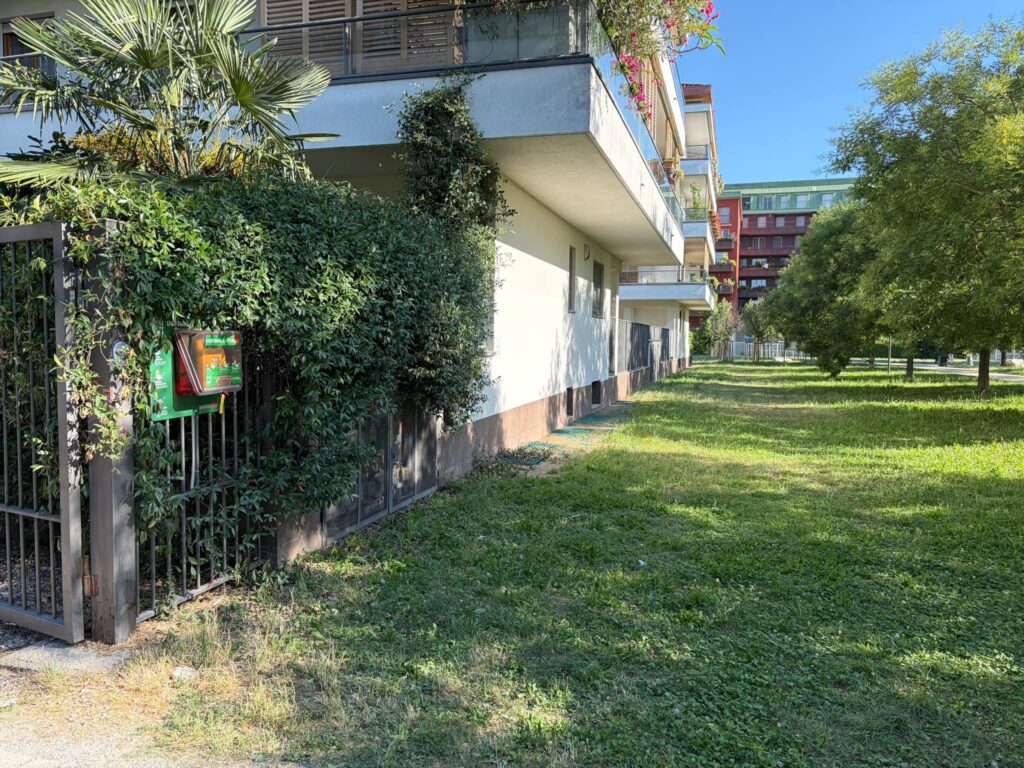 Teca verde salvavita per defibrillatore DAE da esterni installata a ridosso di una siepe in un'area comune residenziale.